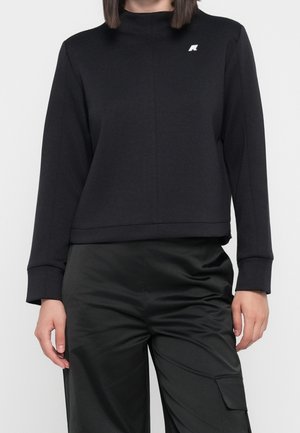 Vrouw draagt een zwart sweatshirt met lange mouwen met een klein wit logo op de borst en zwarte satijnen broek met een zijzak.