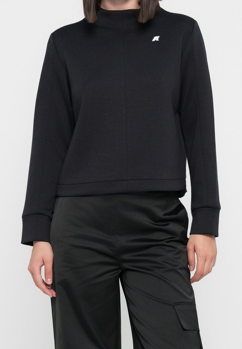 Femme portant un sweat-shirt noir à manches longues avec un petit logo blanc sur la poitrine et un pantalon noir en satin avec une poche latérale.