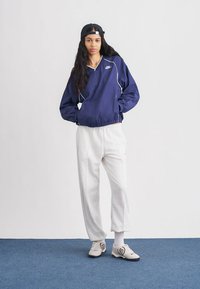 Blusão corta-vento azul-marinho com acabamento branco, punhos e bainha elásticos, combinado com calças de treino cinzentas. O modelo usa um boné preto e ténis de cor clara.