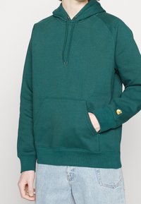 Felpa verde realizzata in materiale morbido. Presenta una tasca frontale, cappuccio con lacci e un accento giallo del logo sulla manica. Polsini e orlo a costine.