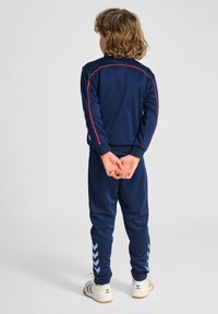 Marineblaues Trainingsanzug-Set mit roten Akzenten, bestehend aus einer Jacke mit Reißverschluss und einer Hose. Die Hose hat hellblaue Zickzackmuster entlang der Seiten.