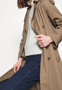 Jasnobrązowy trencz o podwójnym zapięciu, z guzikami i kapturem, noszony na jasnoniebieskim swetrze i ciemnych dżinsach.