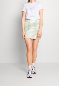 Bílé tričko s grafickým designem, doplněné krátkou sukní zeleno-bílé kostky se bočním rozparkem. K outfitu se hodí sportovní boty.