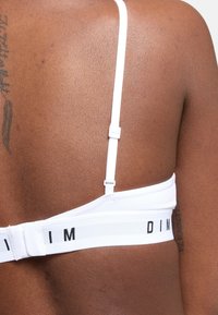 Sujetador deportivo blanco con tirantes ajustables y banda ancha que presenta el nombre de la marca en letras negras. Tejido suave y elástico con un diseño minimalista.