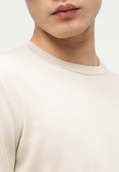 Κοντινό πλάνο στο κάτω μέρος του προσώπου και του λαιμού ενός ατόμου που φοράει ανοιχτό μπεζ πουλόβερ με στρογγυλή λαιμόκοψη σε απλό φόντο.