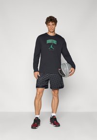 Sort langærmet skjorte med "BOSTON" grafik i grøn, parret med sorte mønstrede shorts og sorte og røde basketballsko, mens der holdes en bold.