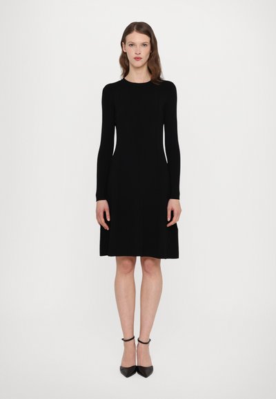 Robe noire à manches longues avec un col rond, corsage ajusté et ourlet évasé. Portée avec des talons ankle-strap noirs. Texture lisse.