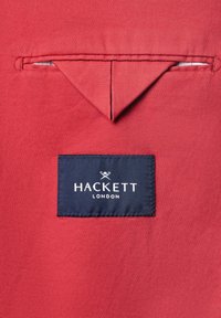 Red fabric with stitched triangular flap above a narrow horizontal pocket, featuring a navy label reading "Hackett London" in white text.