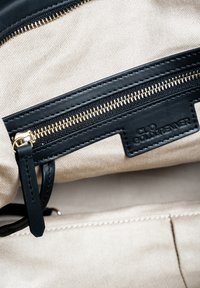 Zwart leren tas interieur met een gouden rits en een beige canvas voering. Bevat een stoffen label met "Clio Goldbrener" in reliëf.