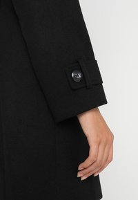 Svart ullkappa med ärm som har en knappstängning och subtila sömdetaljer. Tyget har en mjuk textur och en strukturerad design.