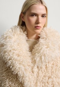 Faux-Fur-Oberbekleidung in Creme mit einer lockigen Textur, über einem beigen Strickpullover geschichtet. Weiche, flauschige Kragenverzierung sichtbar.