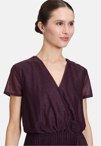 Robe portefeuille violette avec un décolleté en V profond et des manches courtes. Le tissu a un léger éclat et une taille froncée avec une jupe plissée.