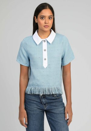 Femme portant un haut à manches courtes bleu clair avec col blanc, trois boutons décoratifs et ourlet à franges, assorti à un jean bleu.