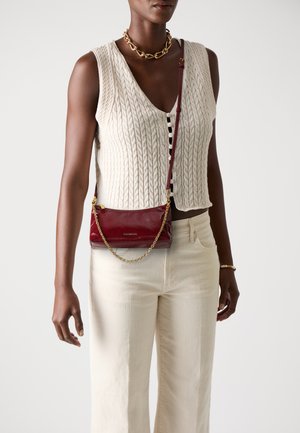 Red leather crossbody bag with a gold chain strap, featuring a smooth texture and subtle branding. Worn over a cream, knitted top.