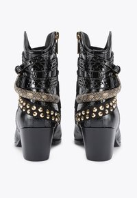 Bottines à talons noirs présentant un matériau texturé, des sangles croisées, des accents métalliques et des clous dorés sur le côté.