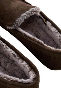 Pantofole in suede marrone con fodera in pelliccia, caratterizzate da una consistenza morbida e una forma arrotondata. Accenti di pelliccia grigia lungo l'apertura.