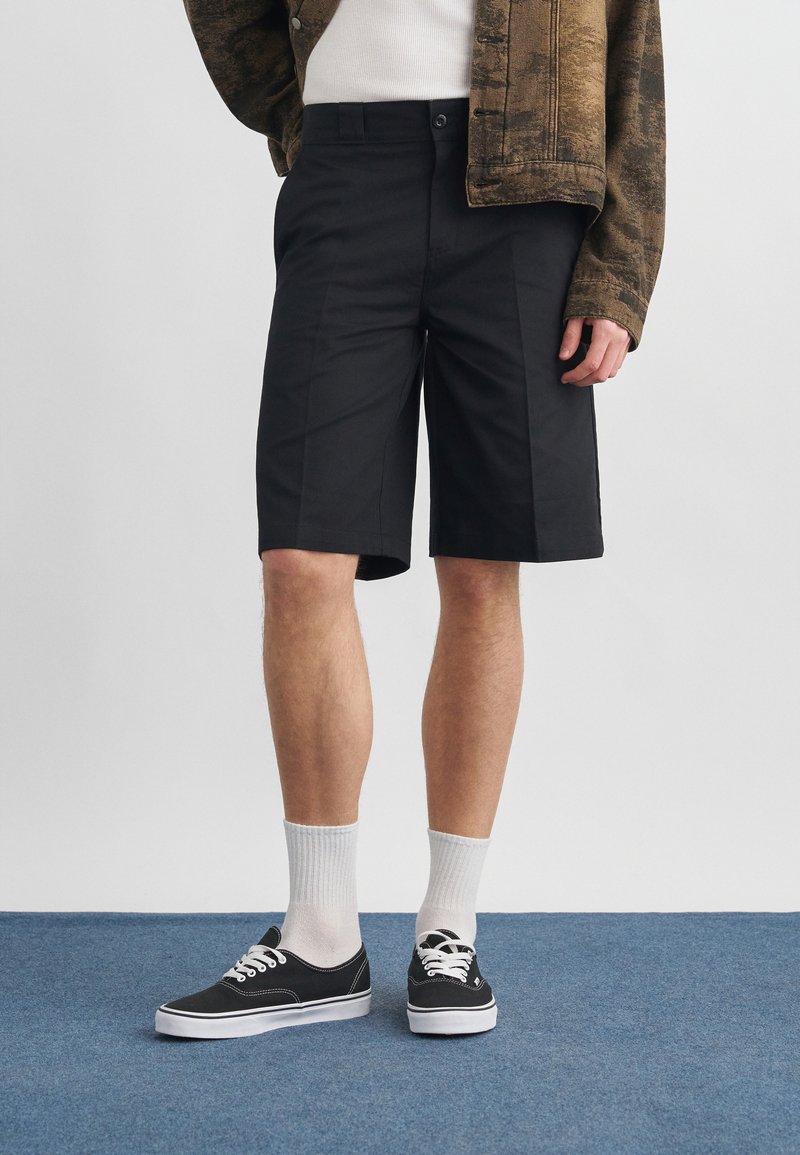 Uomo che indossa pantaloncini neri fino al ginocchio, calzini bianchi, scarpe da ginnastica nere, camicia bianca e giacca marrone a motivi, in piedi su un pavimento blu.