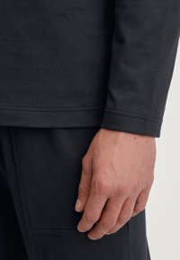 Donkergrijze lange mouw shirt met ribbelmanchetten, met een afgeronde zoom en een gladde textuur. Een hand is zichtbaar nabij de onderste zoom.