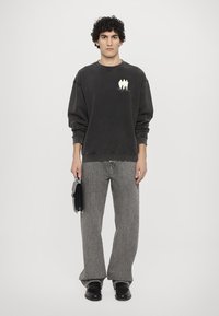 Svart hettegenser med rund hals og hvit grafikk på brystet, kombinert med grå vide jeans og svarte sko, holder en liten svart veske.