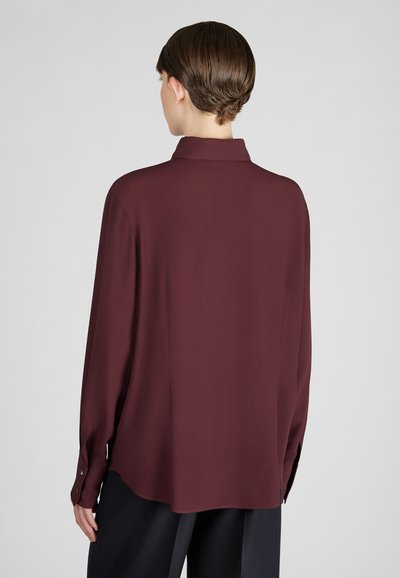 Persoon gezien van achteren, gekleed in een langzichtig, donker bordeauxrood knoopshirt en losse zwarte broek tegen een neutrale achtergrond.