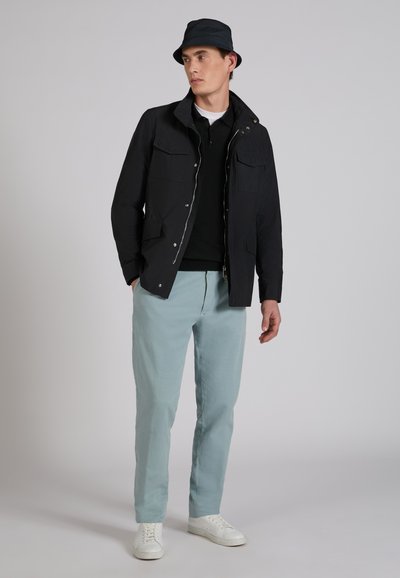 Mężczyzna w czarnym kapeluszu typu bucket, czarnej kurtce na czarnej koszulce, jasnoniebieskich spodniach i białych trampkach, stojący na tle szarego tła.