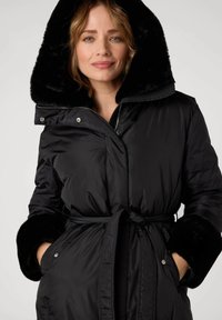 Manteau rembourré noir avec une capuche et des poignets en fausse fourrure, fermeture éclair, ceinture réglable avec cordon de serrage, poches latérales et texture lisse et brillante.