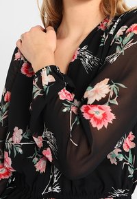 Donna che indossa una blouse nera a fiori con fiori rosa e bianchi, che si tiene la spalla sinistra con la mano destra.