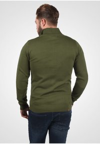 Grüner gestrickter Pullover mit hohem Kragen, gerippten Bündchen und Saum, ausgestattet mit strukturierten Ellenbogenpatches, getragen über dunkelblauen Jeans.
