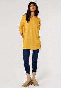Pull oversized jaune en matériau doux, avec des manches larges et une coupe décontractée, associé à un jean skinny bleu marine et des bottines beige clair.