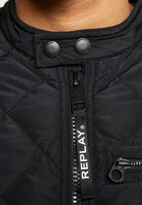 Schwarze gesteppte Jacke mit hohem Kragen, schwarzen Druckknöpfen und sichtbarem Frontreißverschluss mit Logo-Anhänger. Strukturiertem Stoff mit detaillierter Nahtführung.