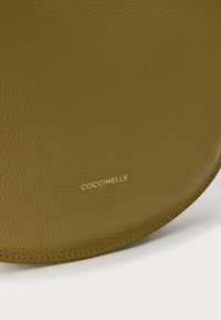 Zielona oliwkowa torba ze skóry o fakturowanej powierzchni; posiada zaokrąglony kształt i złote logo „Coccinelle” wytłoczone na przodzie.