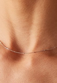 Zilveren ketting met kleine ronde kralen langs de lengte, rustend op de huid. Gladde textuur, delicate ontwerp.