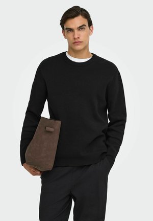 Jeune homme portant un pull noir et un pantalon sombre, tenant une grande pochette en daim marron sur un fond uni clair.