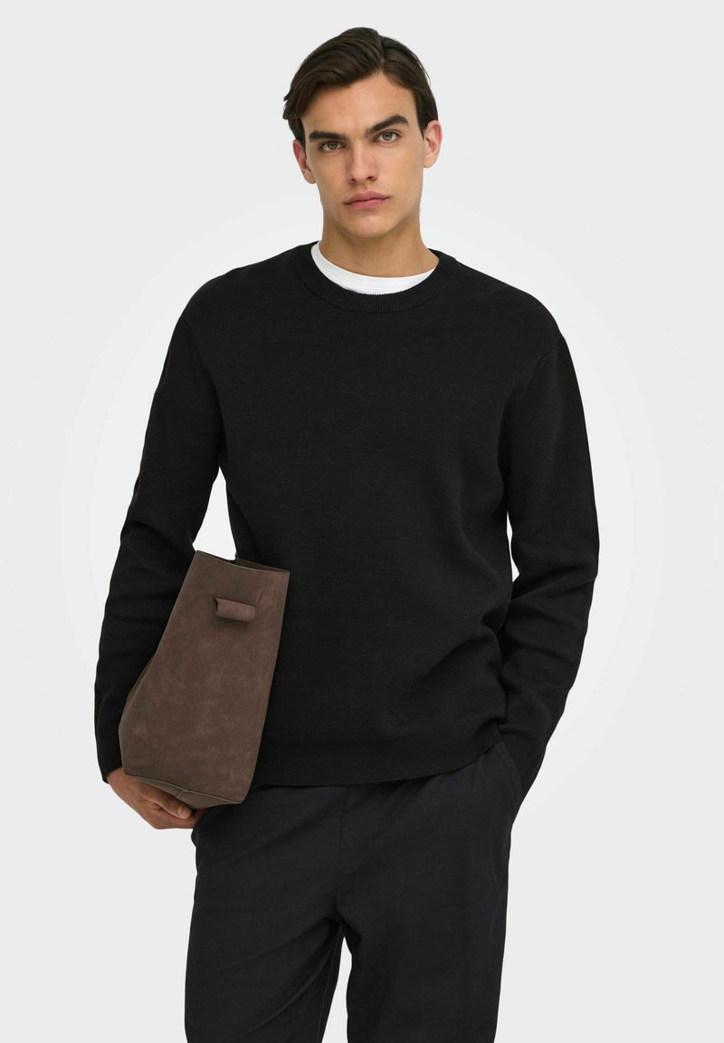 Jeune homme portant un pull noir et un pantalon sombre, tenant une grande pochette en daim marron sur un fond uni clair.
