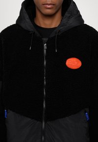 Must have hoodiga musta fliisjakker, millel on tõmblukk, oranž logoäpp ja sinised aktsendid taskutel. Tekstureeritud, soe materjal.