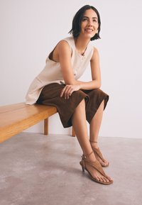 Haut beige sans manches avec boutons, short en suède marron et sandales à talons couleur tan. Modèle assis sur un banc en bois devant un fond neutre.