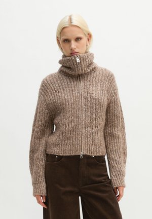Donna bionda che indossa un maglione marrone in maglia grossa con cerniera e collo alto e pantaloni in velluto a coste marrone scuro su sfondo uniforme.