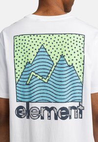 Camiseta blanca con un gráfico de montaña azul, fondo verde con lunares y el texto "element" en la parte inferior en un estilo desgastado.