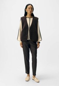 Gilè in pile marrone con apertura frontale, abbinato a una maglietta crema e pantaloni neri. La modella indossa mocassini beige e orecchini semplici. Design minimalista.