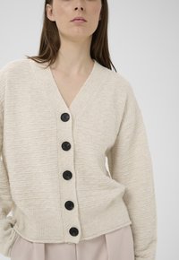 Frau trägt eine strukturierte cremefarbene Strickjacke mit fünf großen schwarzen Knöpfen, kombiniert mit hellrosa Hosen, vor einem schlichten grauen Hintergrund.