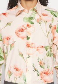 Camisa de botón color crema con patrones de rosas durazno y hojas verdes, llevada por una persona con cabello rizado oscuro.