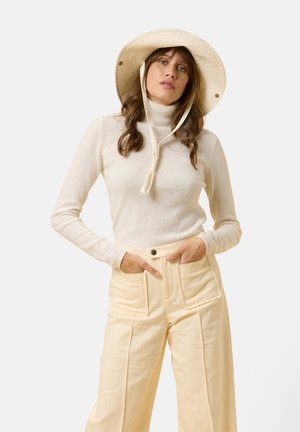 Crème kleur turtleneck trui met lange mouwen, gecombineerd met hoogwarschede lichtgele wijde broek met opvallende voorkeurs. Hoed met brede rand.