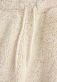 Tessuto in pile color crema con texture morbida e spessa; presenta due cordini bianchi al collo.