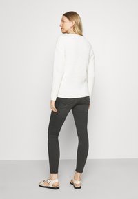 Maglione bianco a costine, jeans skinny grigi scuri e sandali leggeri con dettaglio a fibbia. La vista posteriore mostra una vestibilità rilassata e cuciture minime per il massimo comfort.