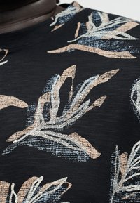 Nahaufnahme eines schwarzen Stoffes mit abstrakten beigen und hellblauen blattähnlichen Mustern auf einer strukturierten Oberfläche.