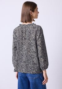 Jeune femme avec des cheveux bruns raides portant un haut à manches longues noir et blanc à fleurs et un jean bleu, tournant le dos contre un fond uni.