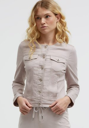 Frau mit langen blonden Haaren, trägt eine hellbeige, geknöpfte Jacke und passende Hose vor einfarbigem Hintergrund.