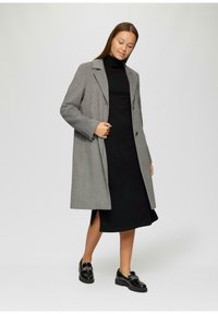 Manteau en laine gris avec une silhouette structurée, revers à cran et deux boutons, porté par-dessus une robe noire, associé à des chaussures noires.
