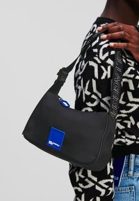 Borsa a spalla nera in nylon con forma curva, toppa del logo blu, tracolla regolabile con branding e chiusura con zip.