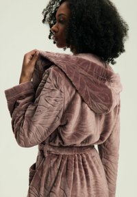 Miękki, teksturowany szlafrok w kolorze mauve z kapturem, ozdobiony wzorem przypominającym liście, z paskiem w talii i bocznymi kieszeniami. Wykonany z pluszowego, przytulnego materiału.